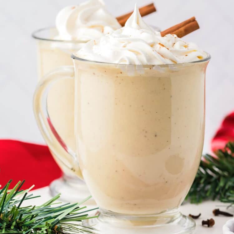 two clear mugs of spiced eggnog topped with whipped cream and cinnamon sticks.