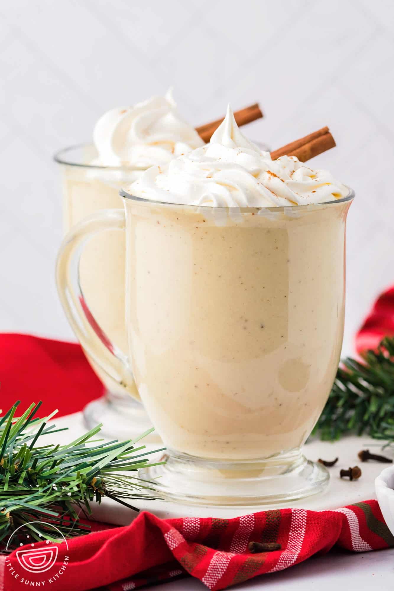 two clear mugs of spiced eggnog topped with whipped cream and cinnamon sticks.