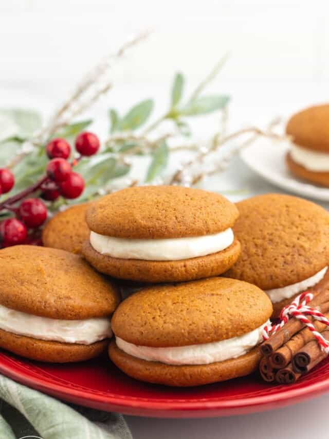 a red plate of gingerbread whoopie pies. A bundle of cinnamon decorates the plate and holiday greenery is in the background
