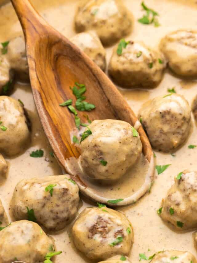 a wooden spoon picking up a meatball from a dish of creamy swedish meatballs.