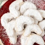 A red plate surrounded by christmas decorations, holding several almond crescent cookies.