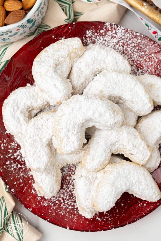 A red plate surrounded by christmas decorations, holding several almond crescent cookies.