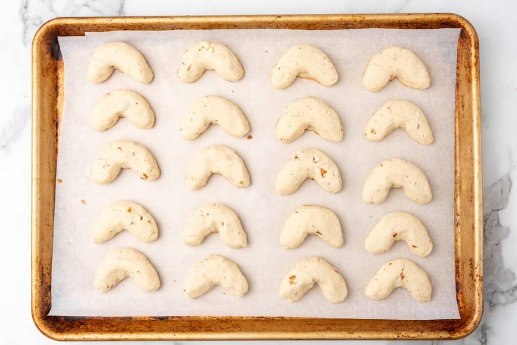 a sheet pan lined with parchment, holding 20 baked almond crescents. The cookies are still very light, not browned on top.