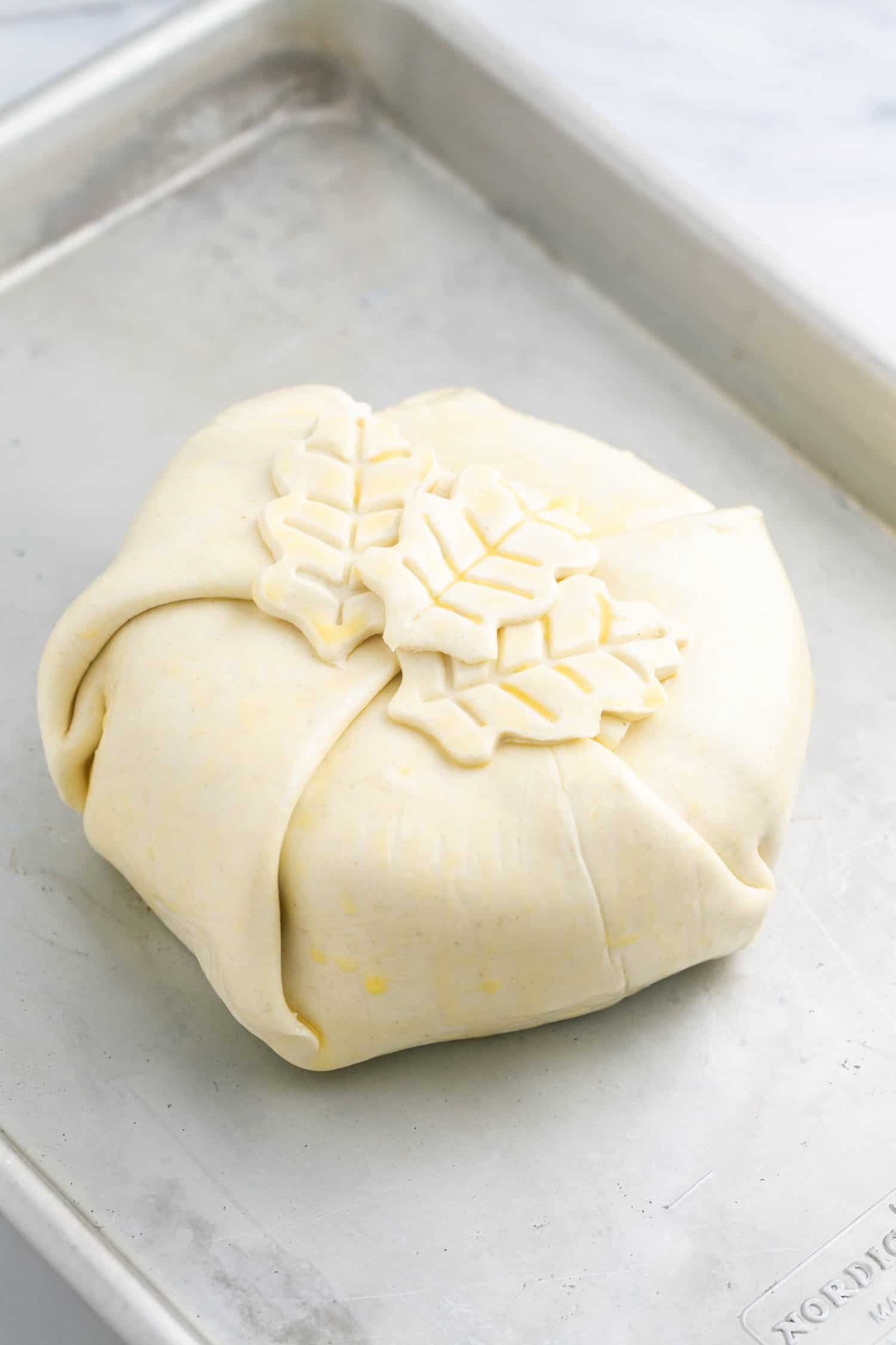 Brie wrapped in puff pastry with leaf cut out shapes, placed on a baking sheet before baking