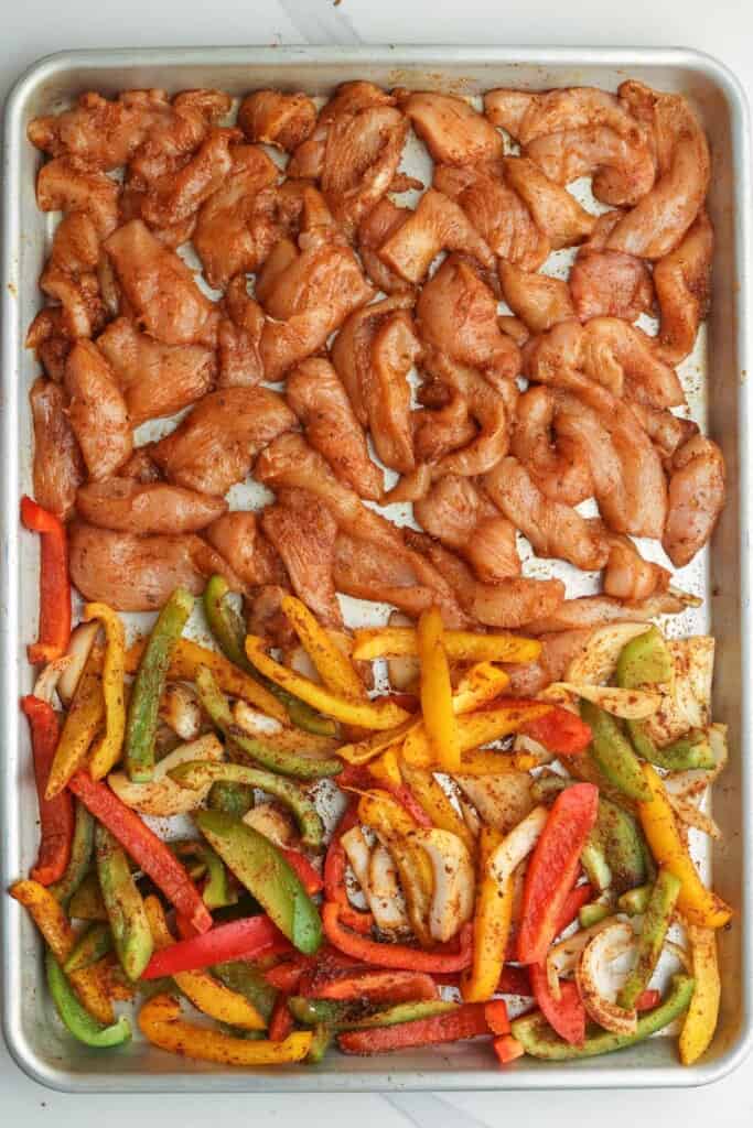 uncooked sliced chicken and veggies, seasoned with fajitas seasonings, on a sheet pan.