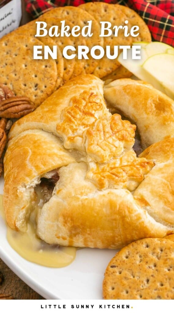 Baked brie in puff pastry served with crackers and thinly sliced apples and overlay text that says "Baked Brie En Croute"