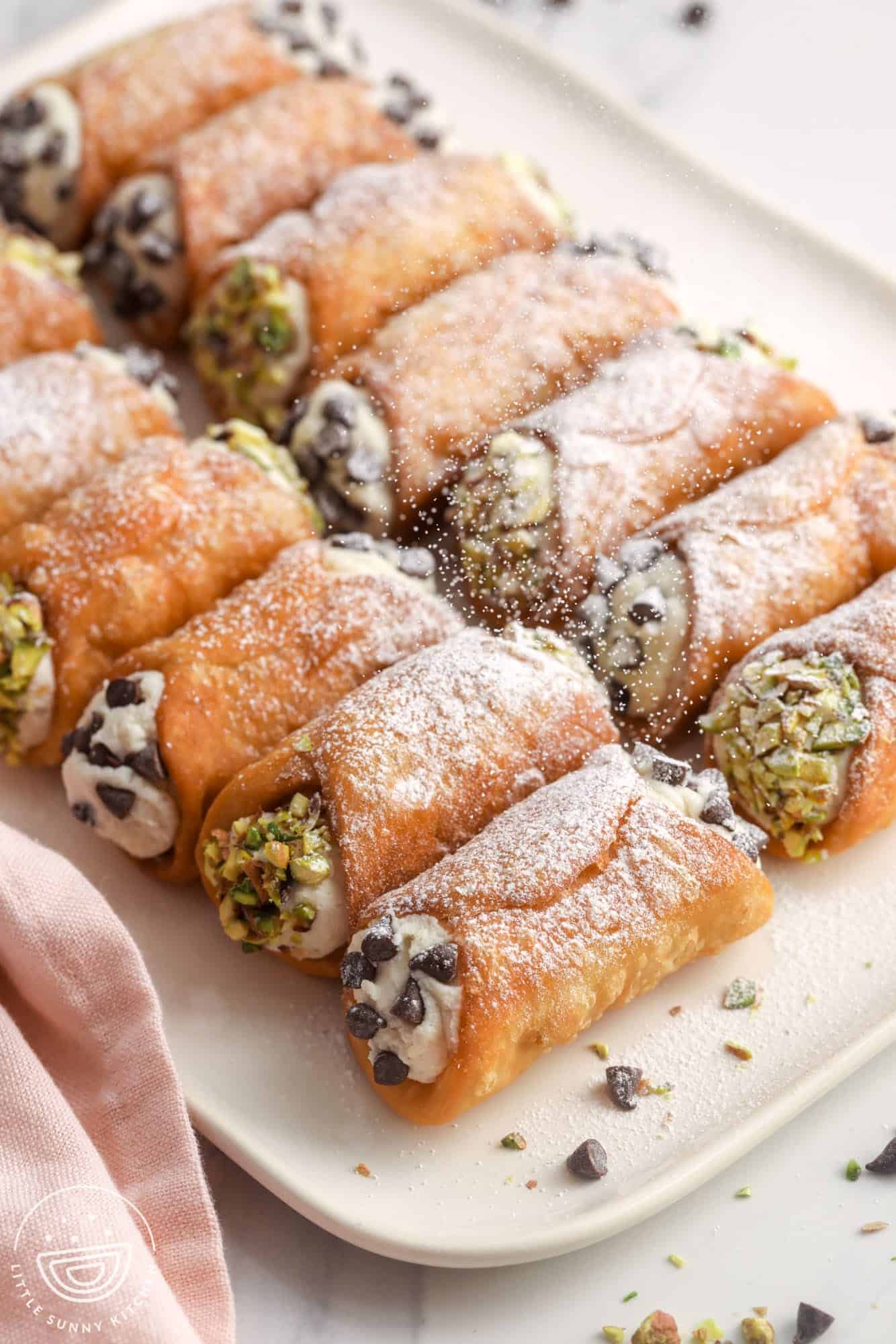 a rectangular white tray holding homemade cannoli filled with creamy filling and dipped in chocolate chips and pistachios. Powdered sugar is being sifted over the cannoli