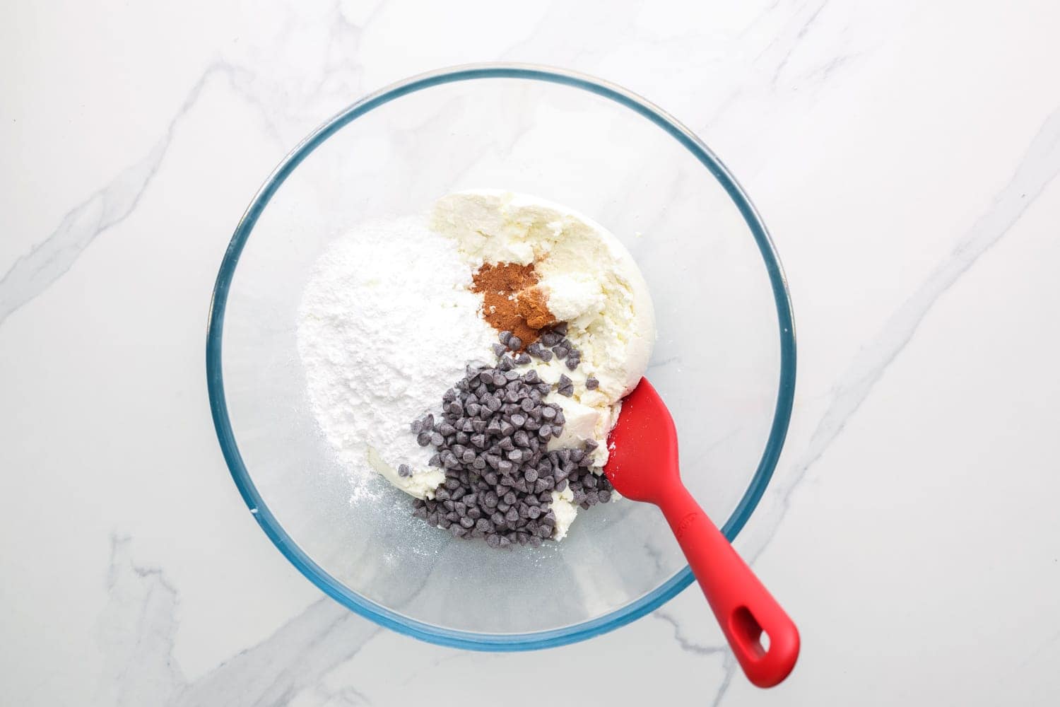 ricotta, sugar, and chocolate chips in a glass mixing bowl with a red spoon. 