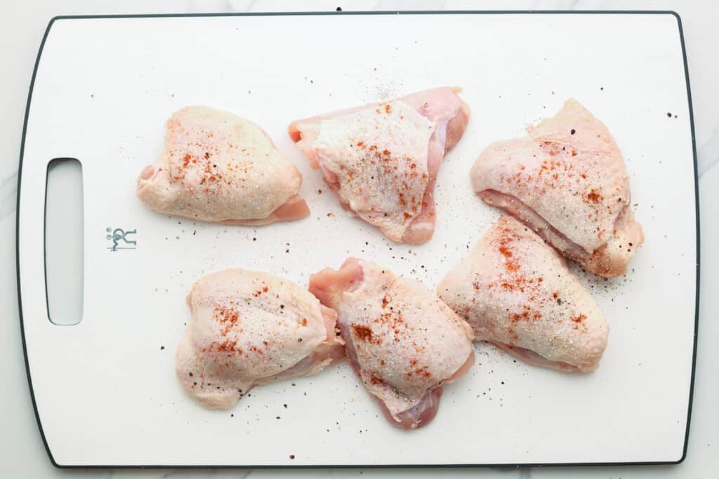 raw chicken thighs with skin on a cutting board, seasoned with salt, pepper, paprika