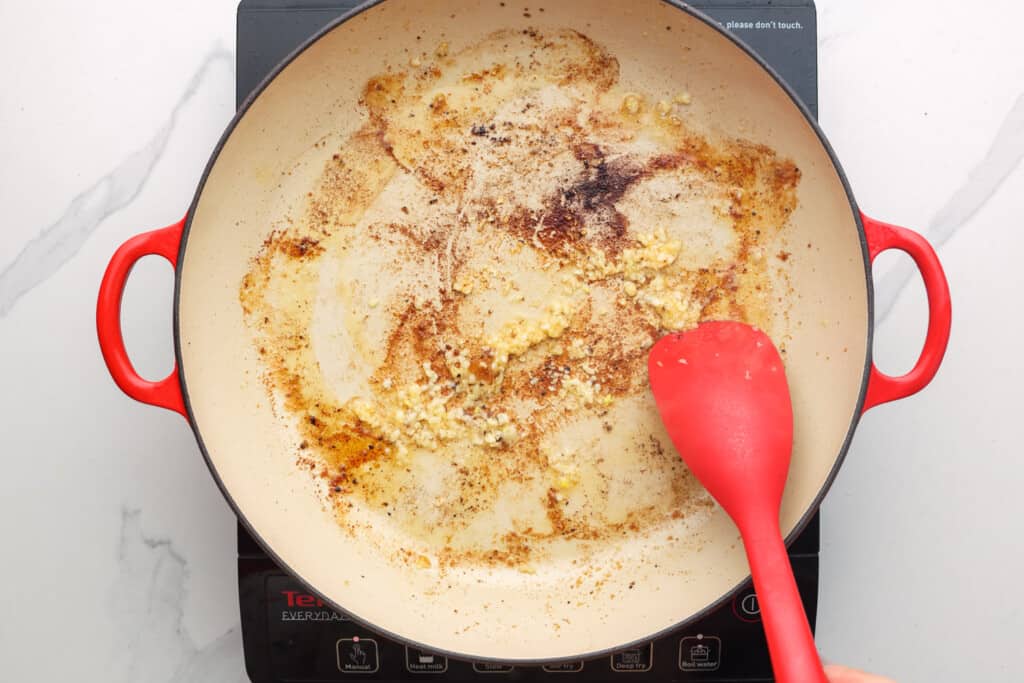 a coated skillet on a burner. In the pan is oil, garlic, and brown bits. A red silicone spatula is scraping the pan.