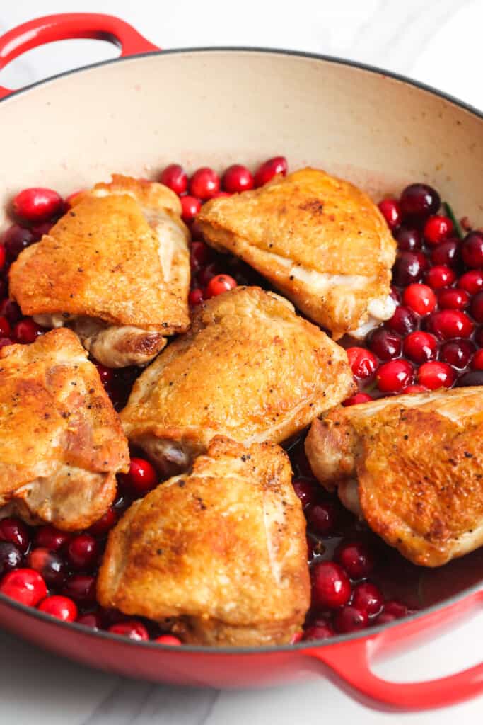 six cooked crispy chicken thighs on top of fresh cranberries in a two handled skillet.
