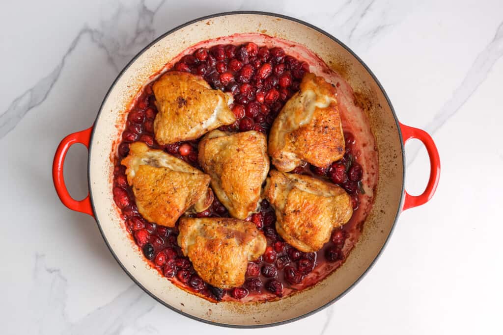 an enameled skillet with two red handles holding six chicken thighs in a cranberry sauce.