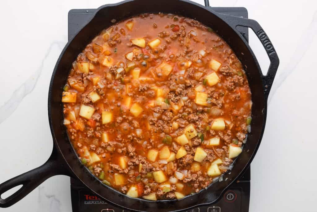 A cast iron skillet simmering potatoes, meat and vegetables to make picadillo