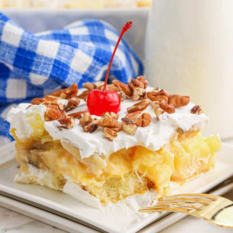 A slice of a no bake twinkie cake topped with whipped topping, pecan pieces, and a maraschino cherry. Served on a square white plate and a golden fork on the side.