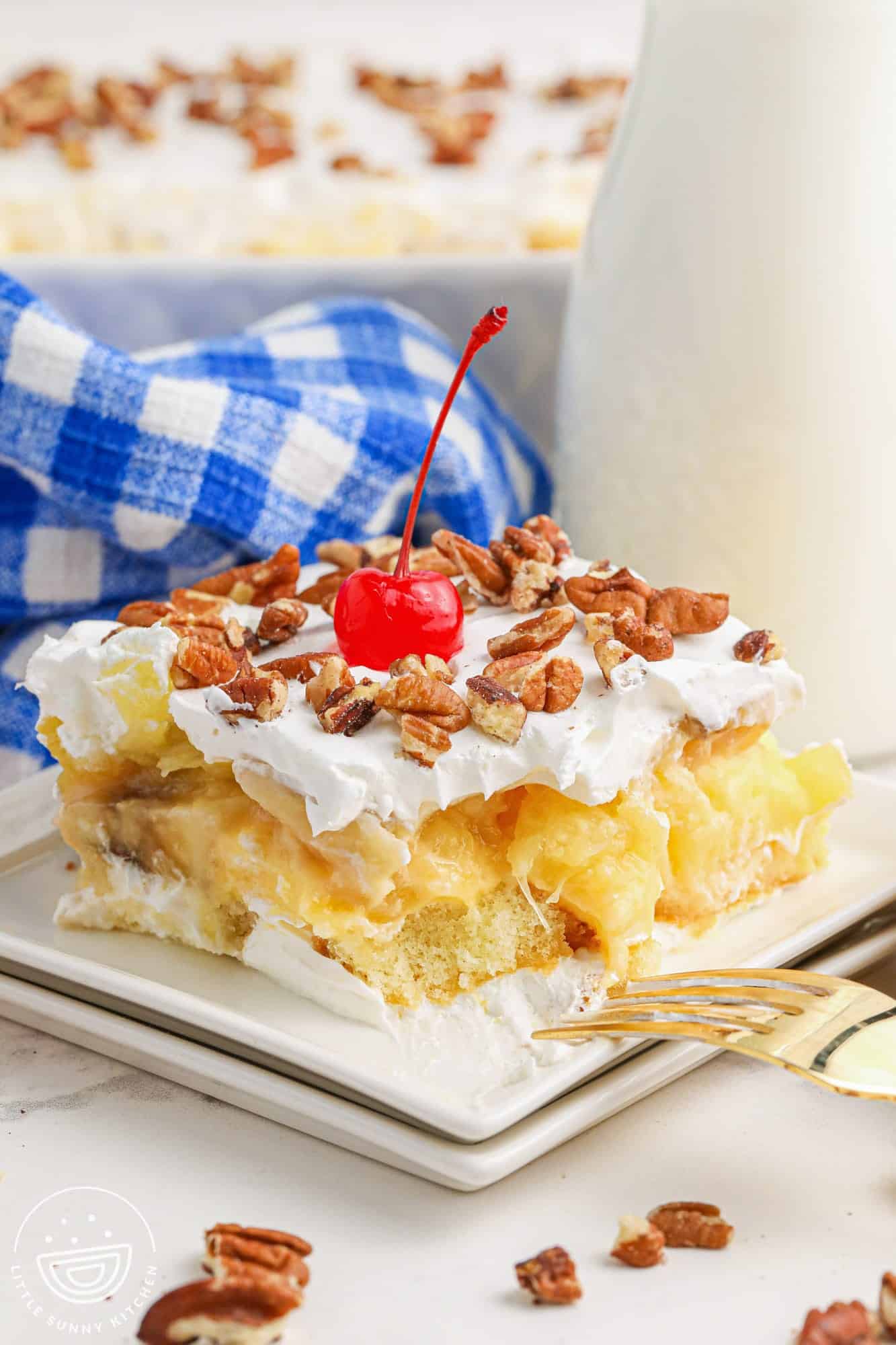 A slice of a no bake twinkie cake topped with whipped topping, pecan pieces, and a maraschino cherry. Served on a square white plate and a golden fork on the side.