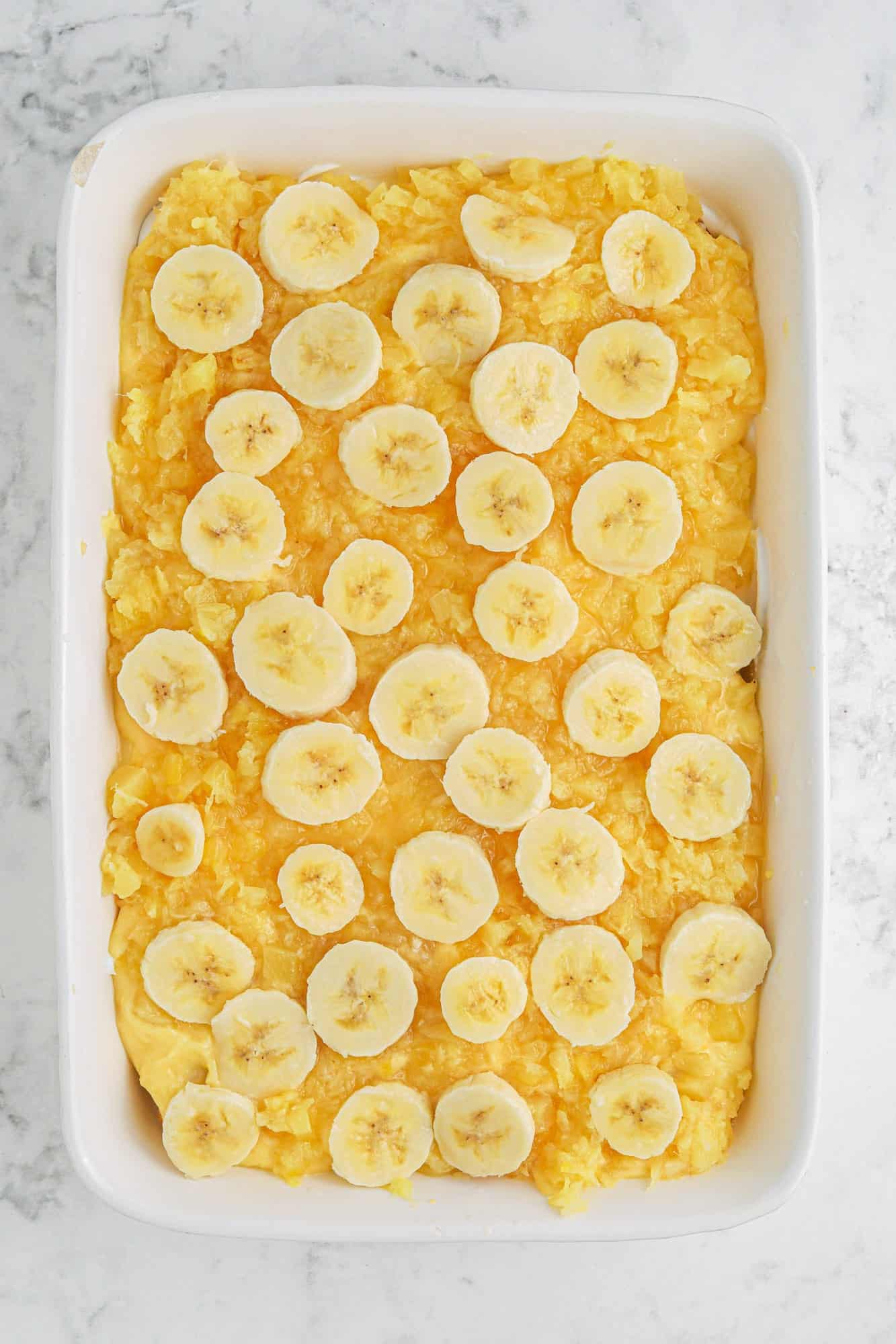 Banana slices layered over crushed pineapple in a 9x13 ceramic white dish