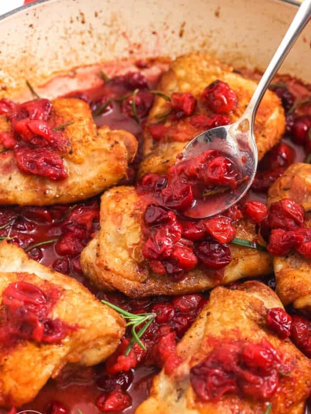 a spoon adding cooked cranberries to the top of cooked chicken thighs in a skillet.