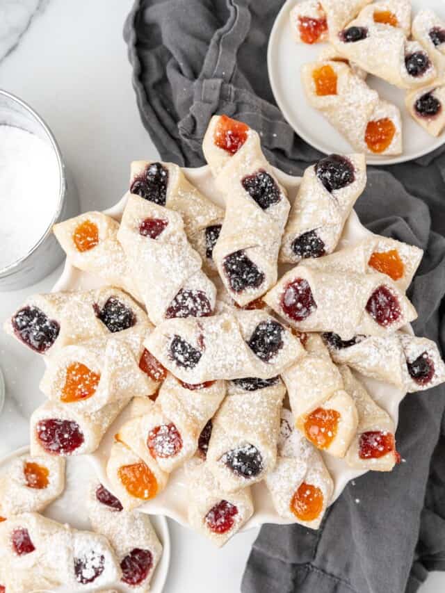 a large platter of polish cookies in the middle of two smaller plates each holding 6 kolaczki. Under the plates is a gray kitchen towel.