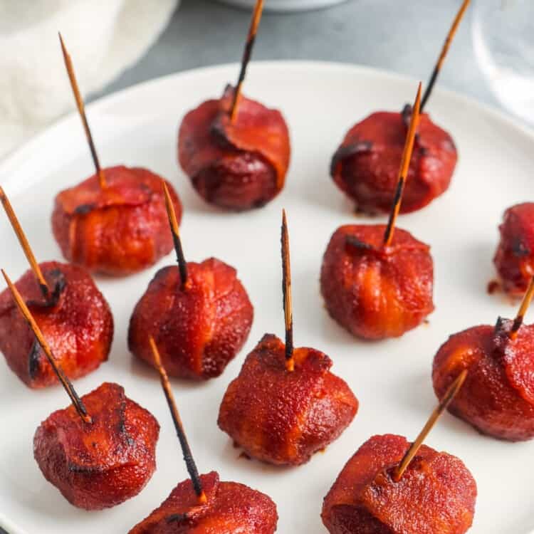 a white plate holding baked chestnuts marinated in red sauce and wrapped with bacon.