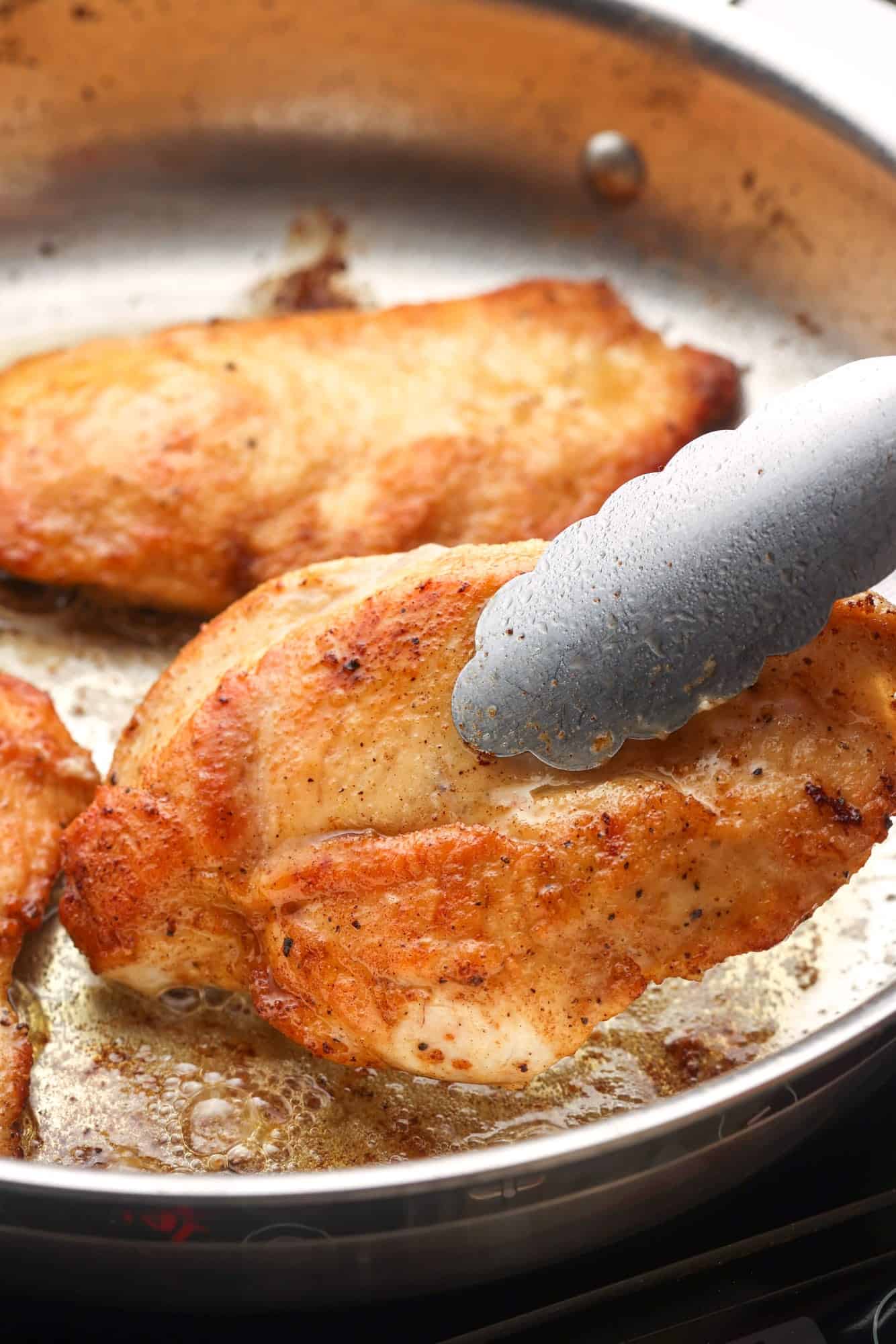 chicken cutlets searing in a stainless steel skillet, one is being flipped with tongs. 