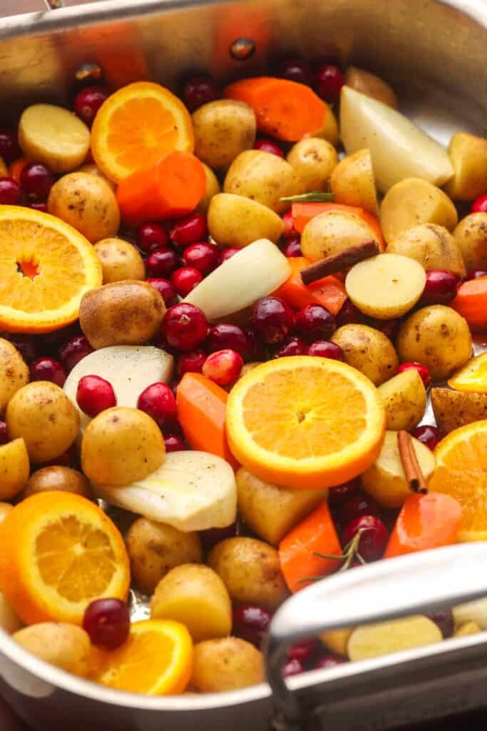 a roasting pan with a mix of cranberries, orange slices, sliced onion, halved baby potatoes, carrot chunks, a cinnamon stick, and fresh herbs.