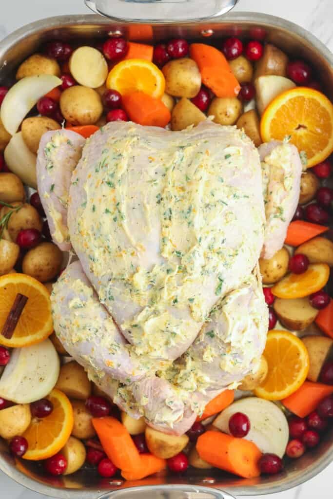 A large roasting pan set up to to into the oven. it's filled with baby potatoes, sliced onion, carrot pieces, cinnamon stick, sliced oranges, and fresh cranberries. A raw whole chicken, covered in seasoned butter is on the vegetables, breast side up
