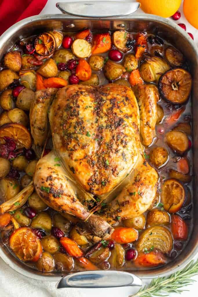 a whole trussed chicken in a roasting pan with a mix of braised onions, carrots, potatoes, and aromatic fruits.
