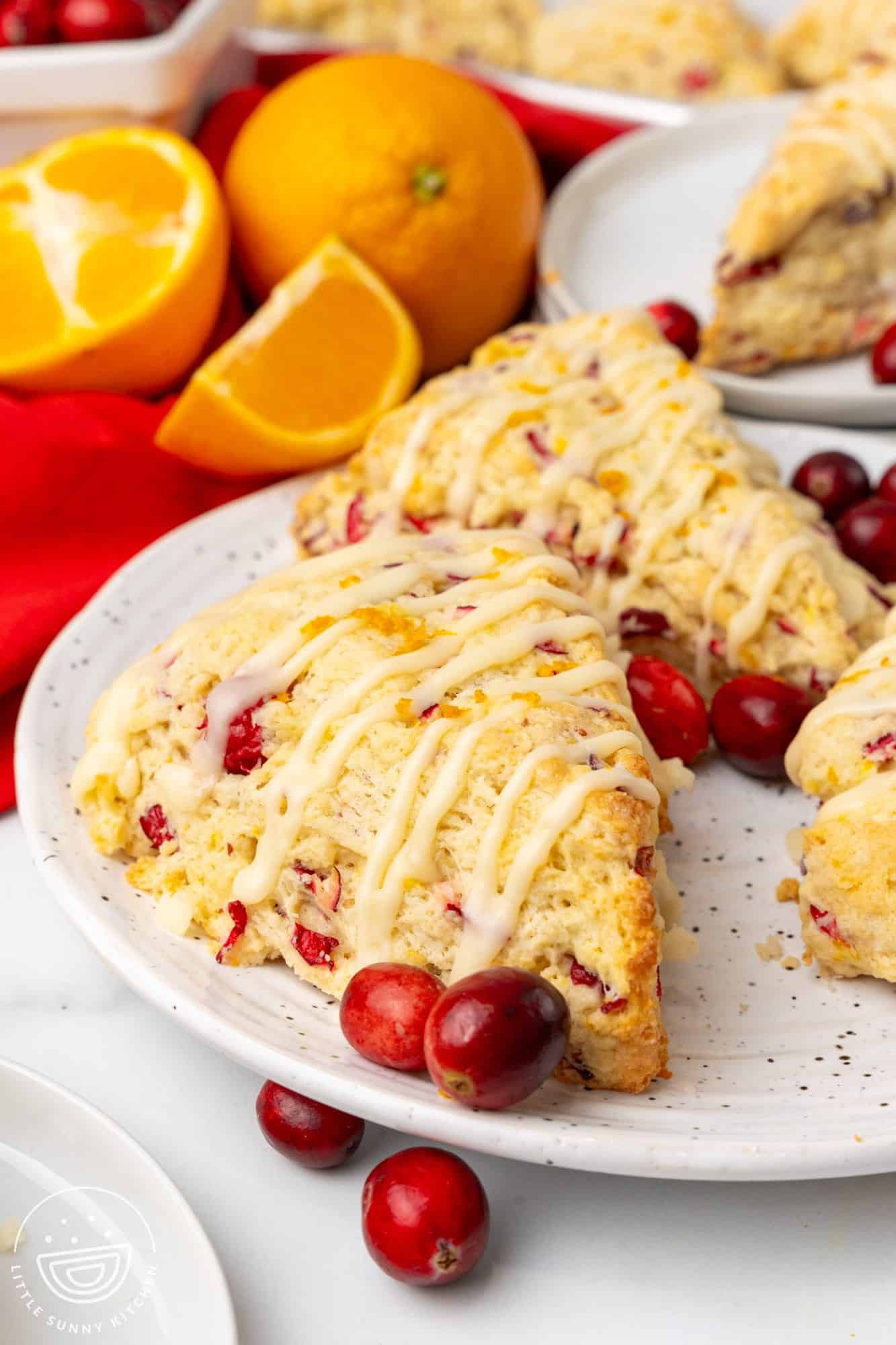 Two cranberry orange scones on a white and gray speckled plate. Fresh cranberries are on the plate and orange wedges are behind it. 