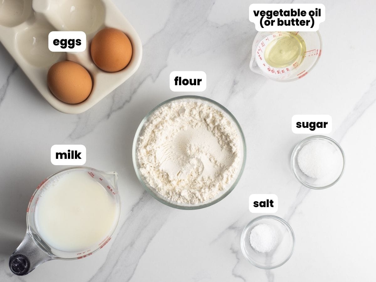 The simple ingredients needed to make crepes: flour, milk, eggs, oil, sugar, salt, all in separate bowls, arranged on a marble countertop.