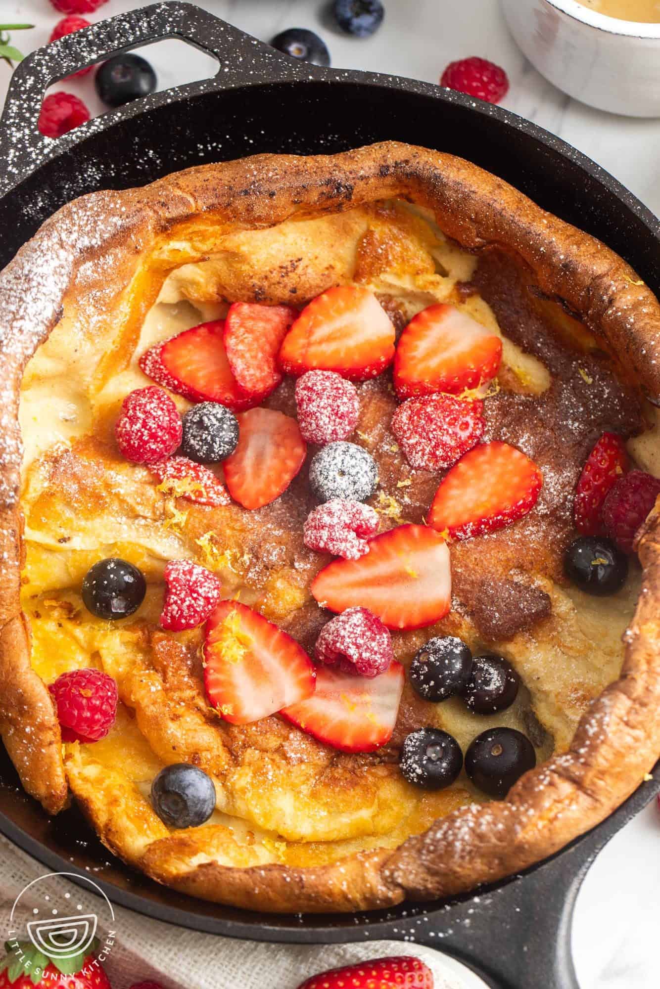 a cast iron skillet holding a german dutch baby pancake topped with powdered sugar, raspberries, blueberries, and sliced strawberries.