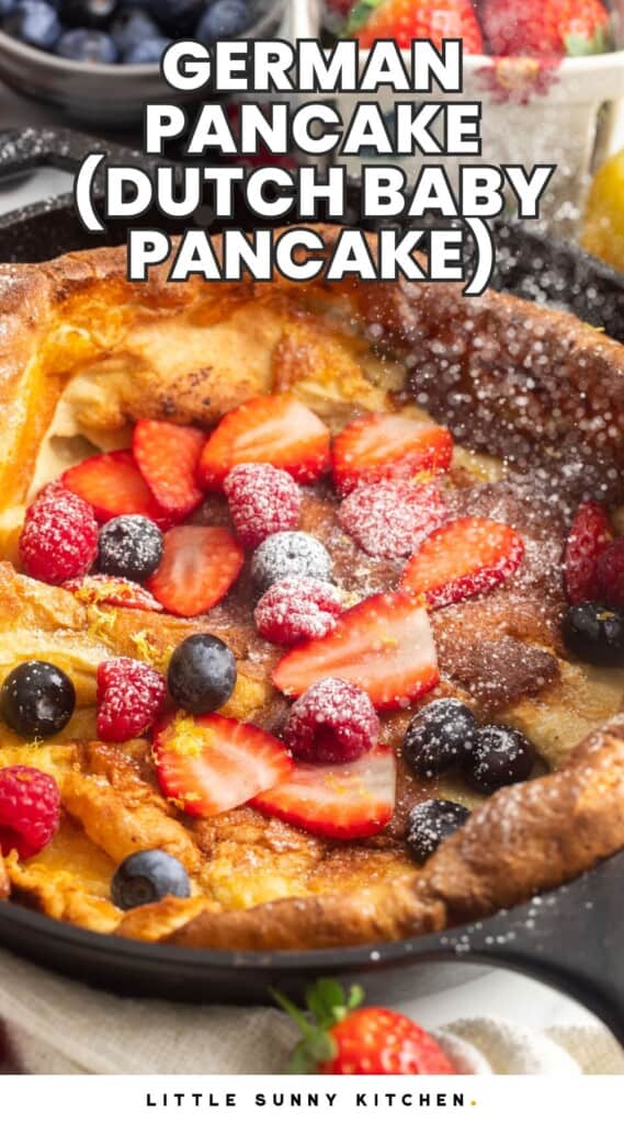 a german pancake in a skillet, topped with fresh berries, sprinkled with powdered sugar. Text overlay says "German Pancake (Dutch Baby Pancake)"