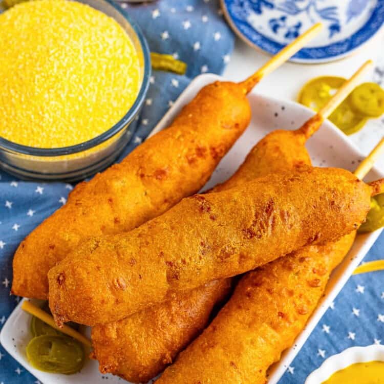 a square plate holding four hand-dipped corndogs with a side of yellow dipping sauce. In the background is shredded cheddar cheese and pickled jalapenos