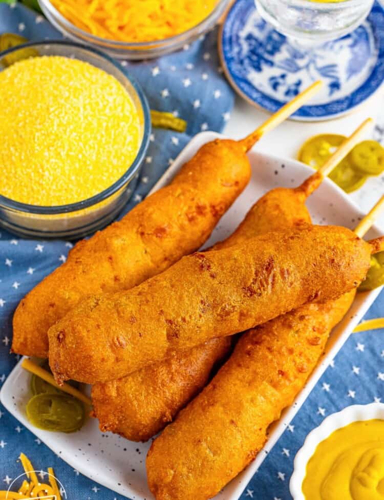 a square plate holding four hand-dipped corndogs with a side of yellow dipping sauce. In the background is shredded cheddar cheese and pickled jalapenos