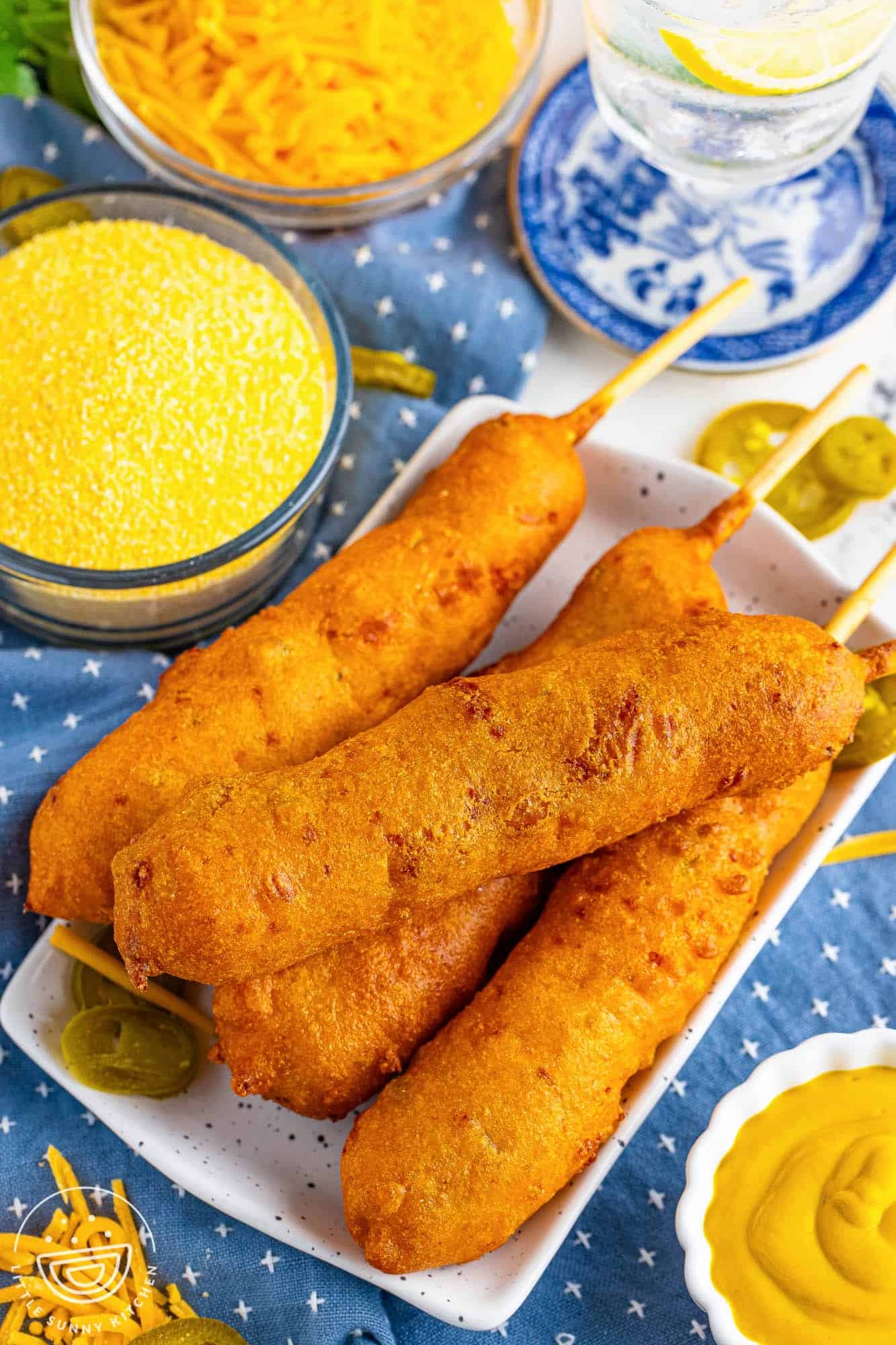 a square plate holding four hand-dipped corn dogs with a side of yellow corn meal. In the background is shredded cheddar cheese and pickled jalapenos