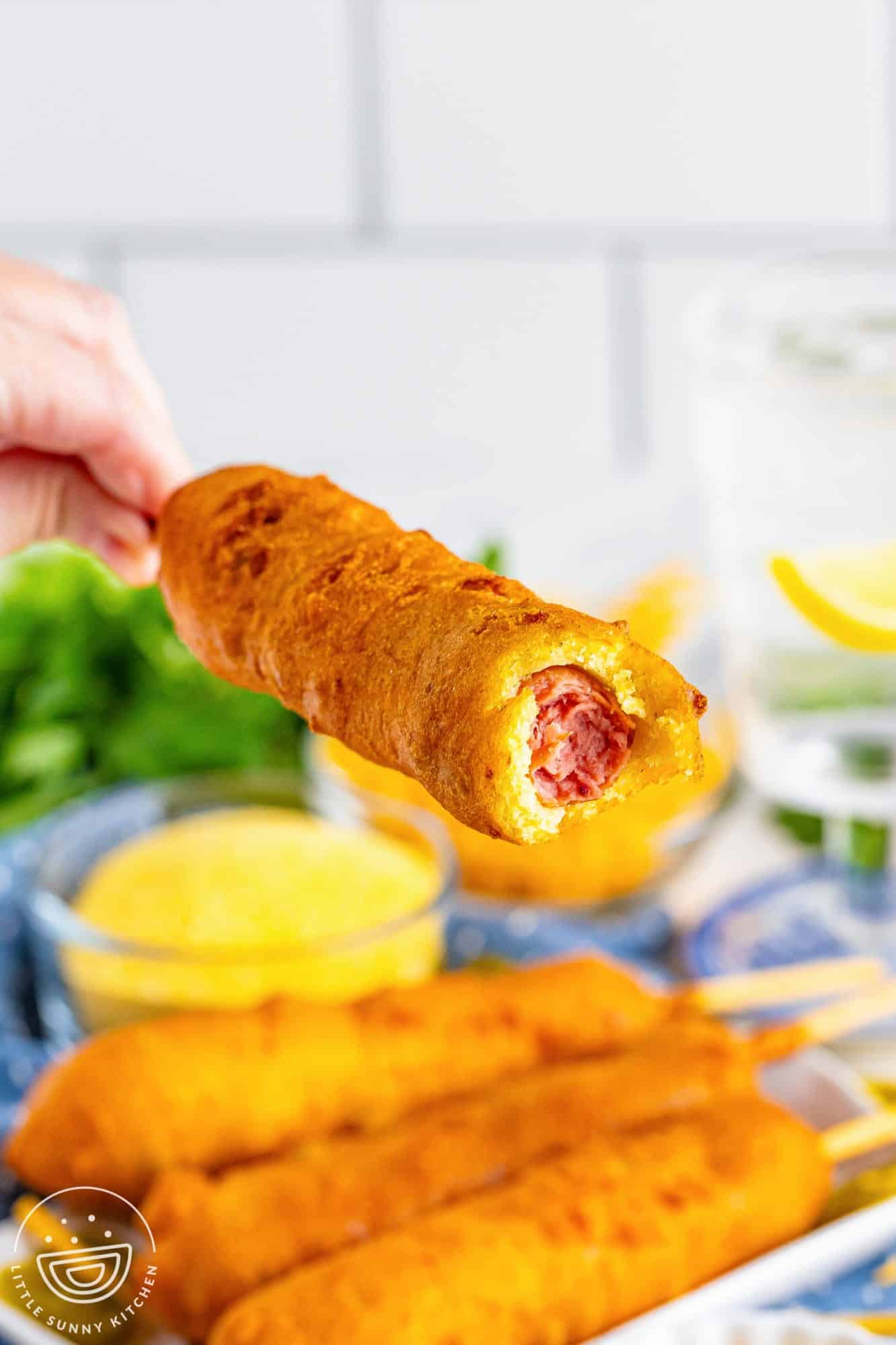 a hand holding a cheddar jalapeno corndog up over a plate of them. A bit has been taken to show what the corn dog looks like inside. 