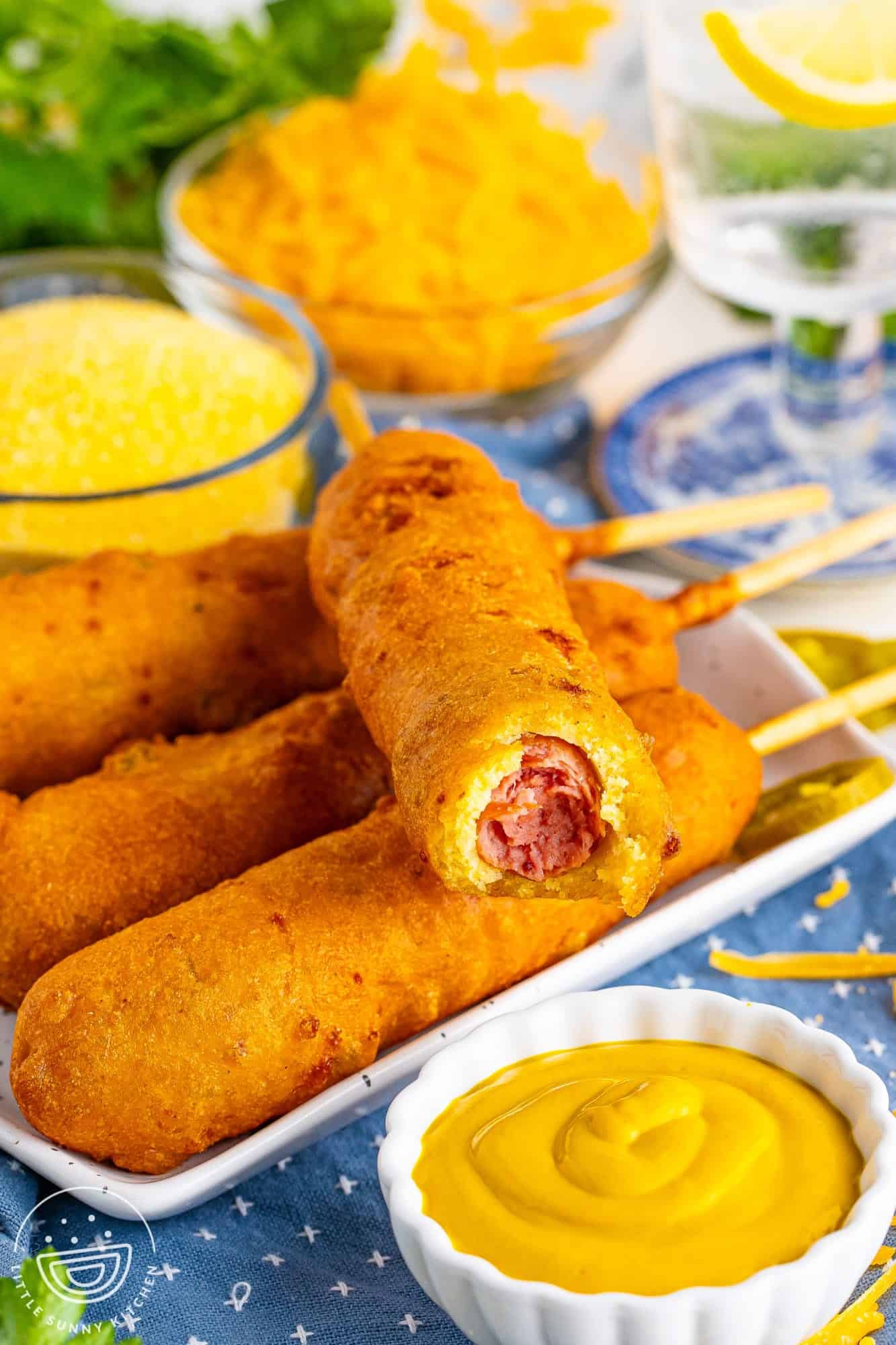 a square plate of four handmade corn dogs with cheddar cheese and jalapenos in the batter. In front of the plate is a small dish of mustard. 