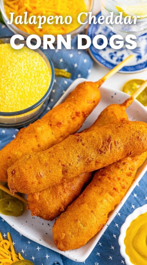 a square plate holding four hand-dipped corndogs with a side of yellow dipping sauce. In the background is shredded cheddar cheese and pickled jalapenos. Text overlay says "jalapeno cheddar corn dogs"