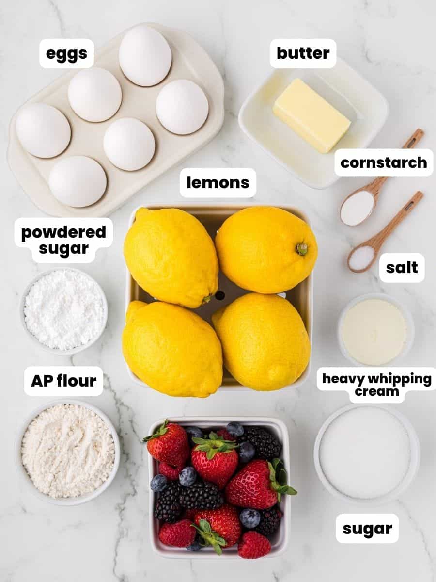 The ingredients for lemon curd tart. In the center is a container of four lemons. Above that are six eggs, a half stick of butter, and spoons holding cornstarch and salt. Below the lemons are bowls holding powdered sugar, whipping cream, granulated sugar, and flour. 