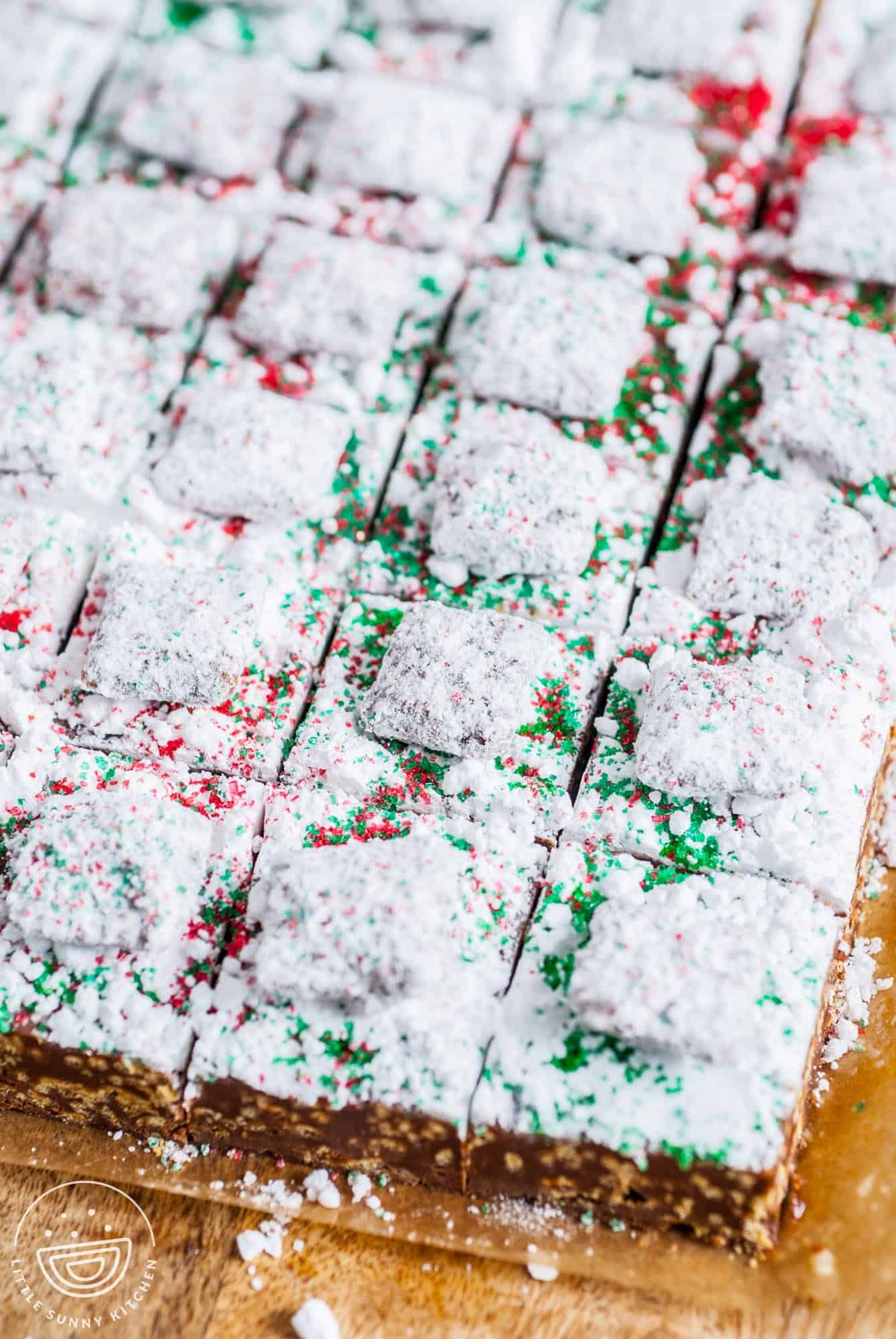 a pan of chocolate crispy fudge topped with powdered sugar, red and green sugar sprinkles, and chex mix muddy buddies. 