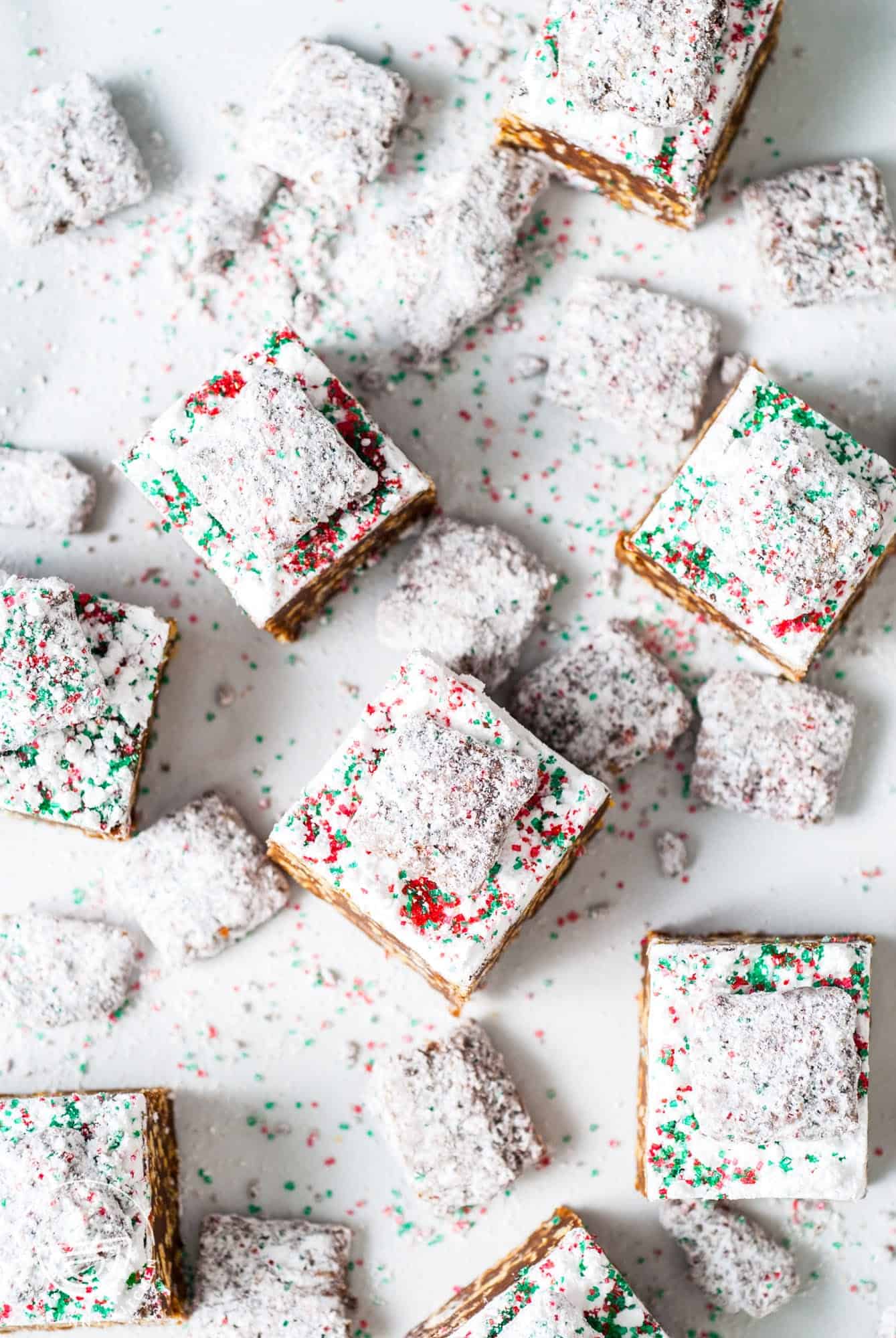 fudge squares on a white surface. The fudge is topped with powdered sugar, muddy buddies, and christmas sprinkles. 