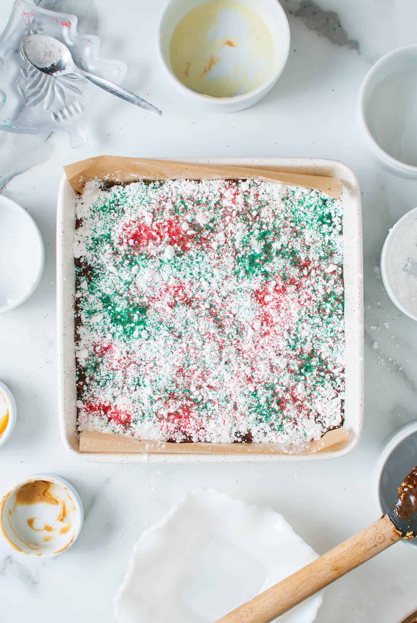 microwave fudge topped with powdered sugar, red and green sugar sprinkles, in a square pan lined with parchment paper. 