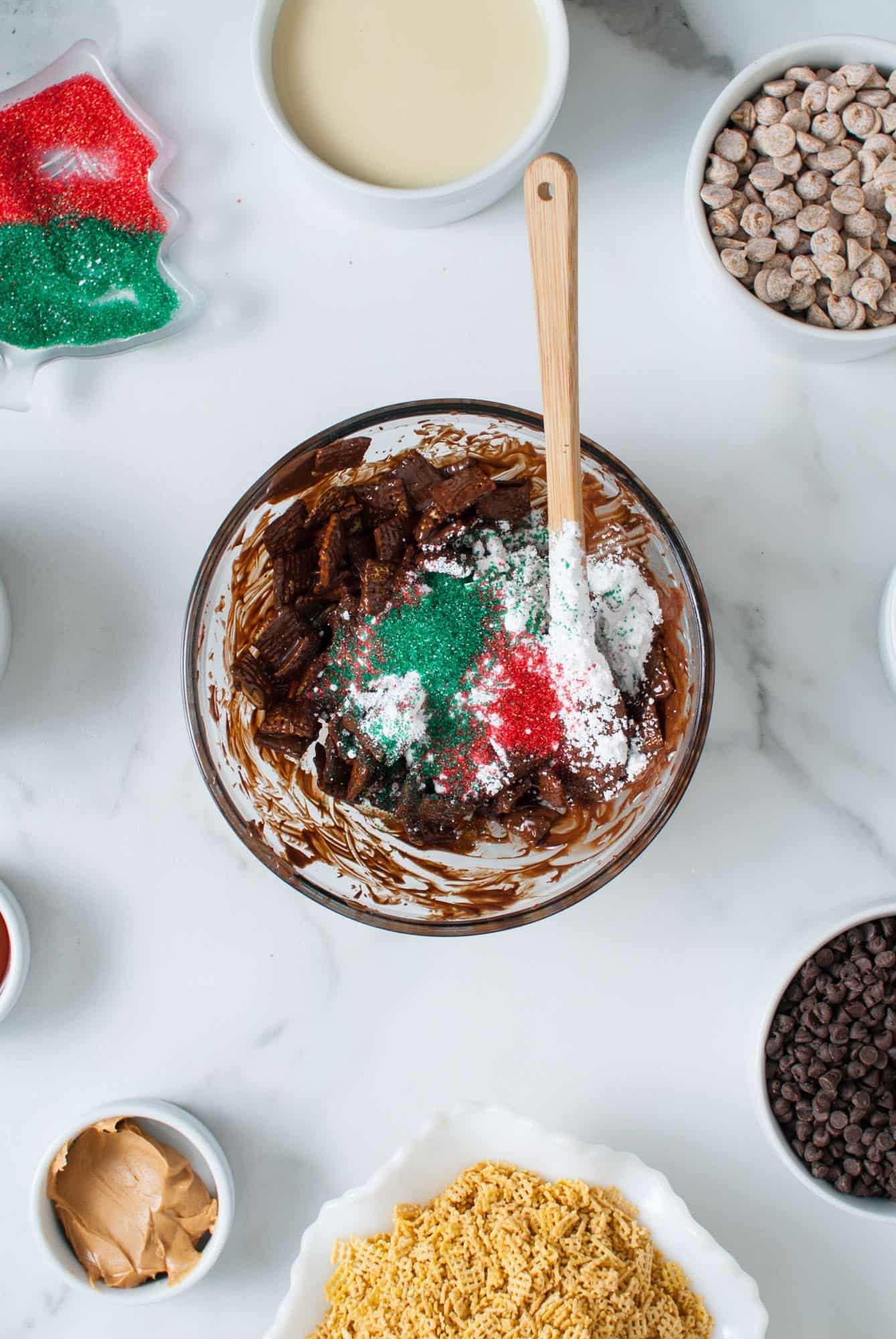 chocolate peanut butter coated chex in a glass mixing bowl with a spatula. Powdered sugar and red and green sprinkles have been added. 