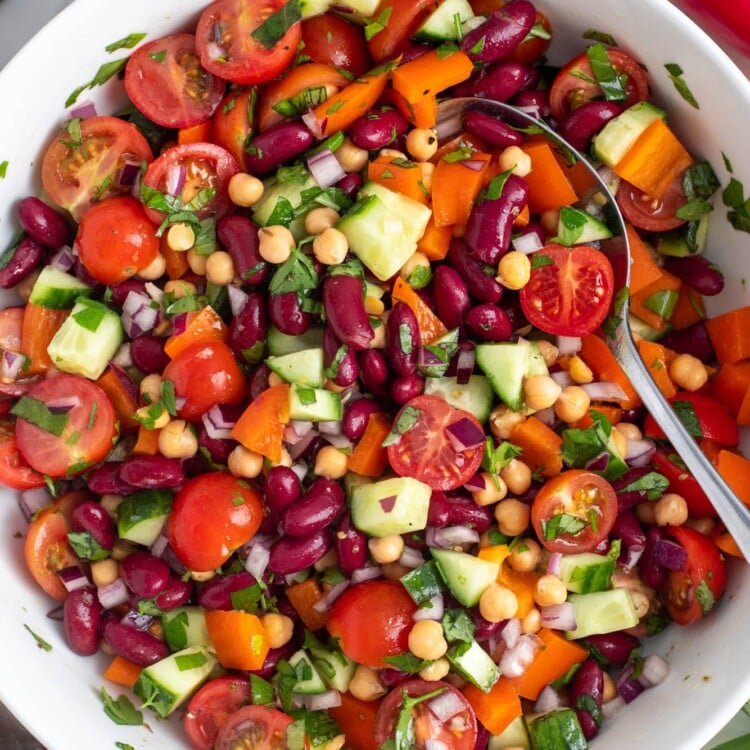 Bean salad with chickpeas, kidney beans, tomatoes, cucumbers, and herbs, in a white serving bowl, viewed from overhead.