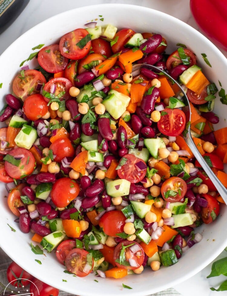 Bean salad with chickpeas, kidney beans, tomatoes, cucumbers, and herbs, in a white serving bowl, viewed from overhead.
