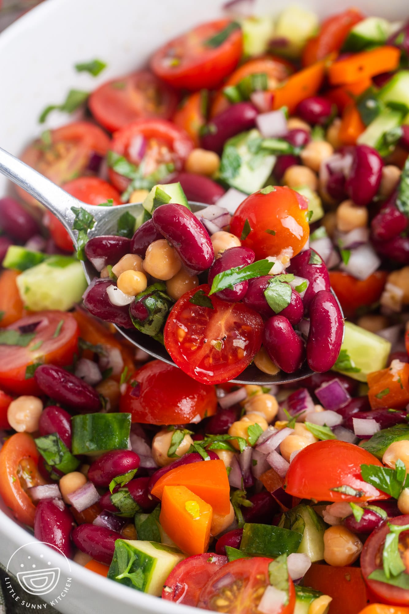 salad with red kidney beans, chickpeas, and vegetables, a scoop held up by a spoon over a bowl of it.