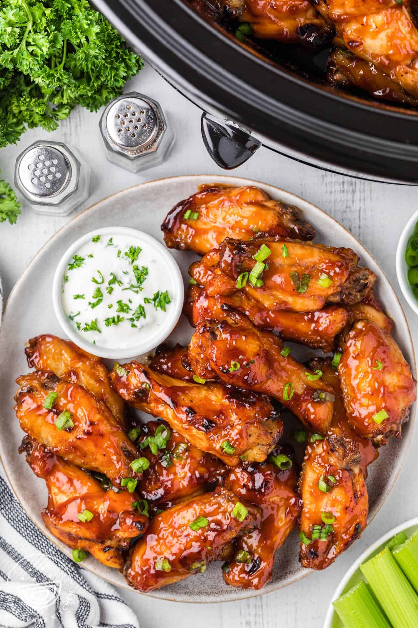 a white plate of bbq chicken wings with a side cup of ranch. This plate is next to a crockpot.