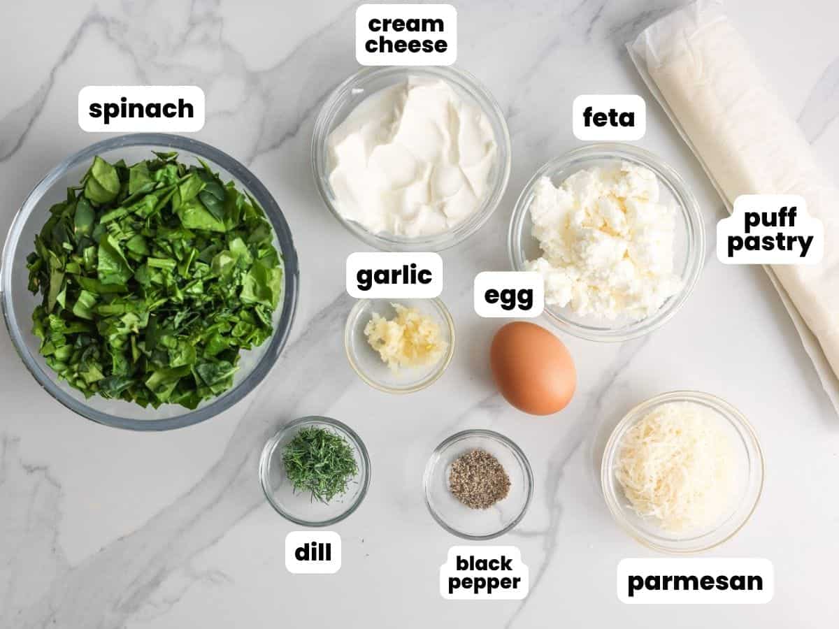 a bowl of fresh chopped spinach next to small bowls of spinach puff ingredients and a package of frozen puff pastry. 