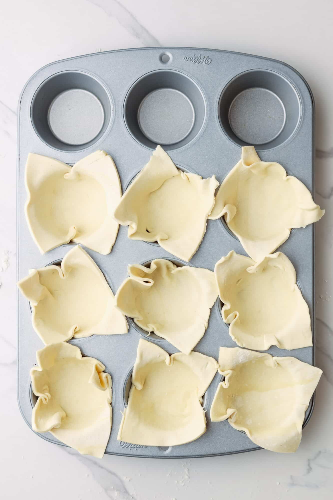 a silver muffin pan. 9 of the spaces are lined with squares of puff pastry dough. 