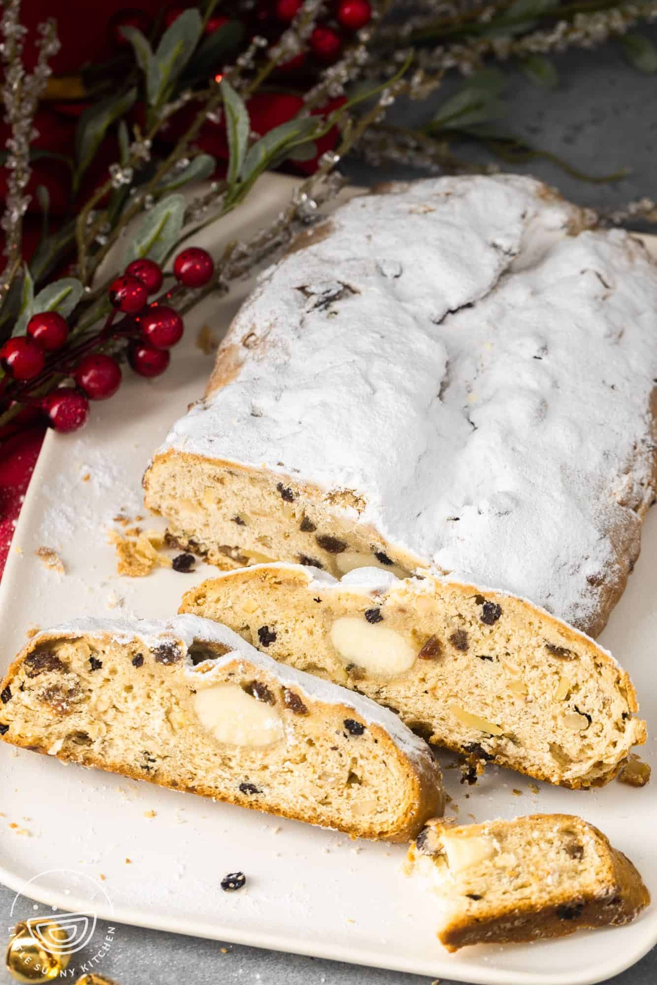 a loaf of stollen bread, dusted heavily with powdered sugar, on a white platter. The loaf is sliced, showing slices with raisins, nuts, and marzipan inside. 