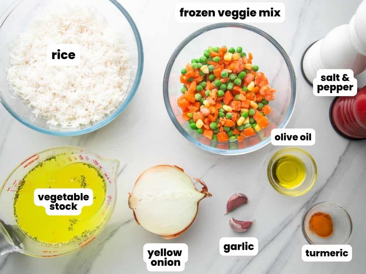 A glass bowl of frozen mixed veggies next to a bowl of rice, a cup of vegetable stock, onion, garlic, olive oil, and turmeric.